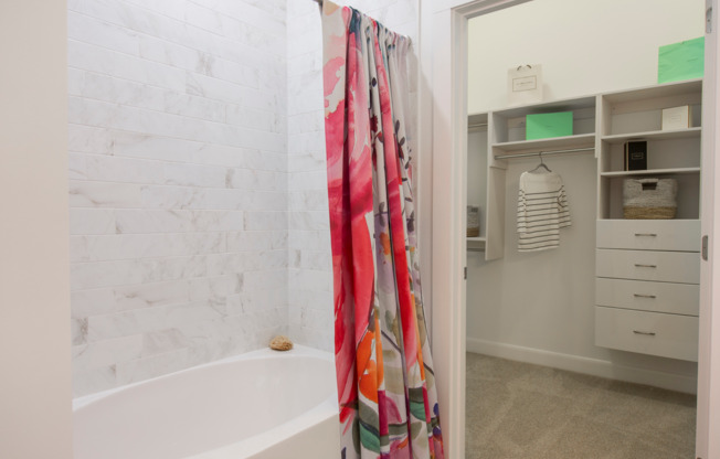 Bathroom with an oval soaking tub, floral shower curtain, and marble tiling to the ceiling. A door leads into a large walk-in closet with beige carpet.