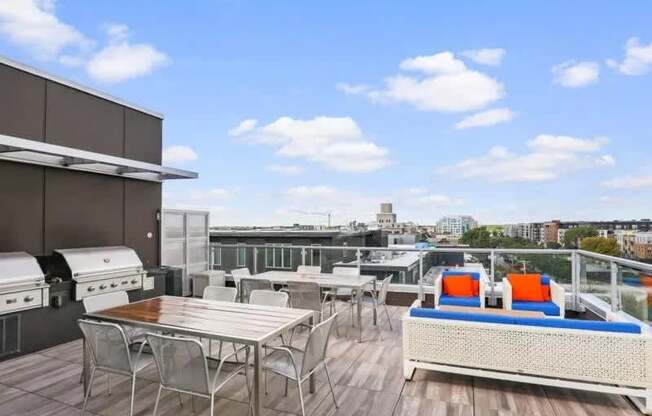 A spacious rooftop gathering area here at Blue and Lime featuring stainless grilling stations, metal dining tables with chairs, and woven lounge seating with blue and orange cushions. Wood-style decking and glass railings frame open views of the surrounding neighborhood, creating an inviting setting for outdoor meals, social gatherings, and relaxed city living.