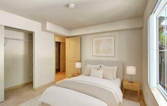 A cozy, well-lit bedroom here at Asbury Park featuring a neatly made bed with white bedding and soft neutral accent pillows, a light upholstered headboard, and matching bedside tables with modern lamps, complemented by plush carpet, an open closet with shelving, and a large window that brings in natural light.