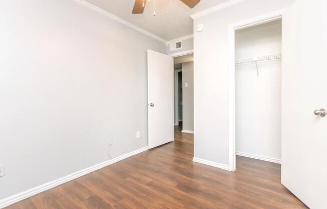 A well-lit room with light gray walls and hardwood flooring. Two doors are visible: one leading to a closet and the other to an adjoining space. A ceiling fan is mounted above, and the room appears clean and organized, with no furniture present.
