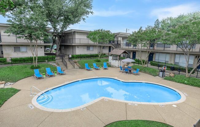 A heart-shaped swimming pool surrounded by green grass and trees. Lounge chairs are arranged around the pool, and a shaded seating area is visible. Nearby buildings feature balconies and a clear blue sky above.
