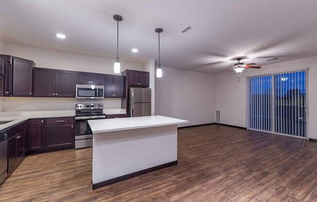 A spacious open-concept kitchen and living area here at The Park featuring dark wood cabinets, a bright island, wood-style floors, and large sliding glass doors with vertical blinds, creating a modern and inviting gathering space.