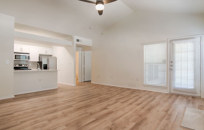 an empty living room with a ceiling fan and a kitchen in the background