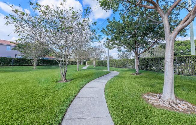 A grassy park with a winding concrete pathway surrounded by small trees and hedges. The landscape features bright green grass under a blue sky with fluffy clouds, creating a peaceful and serene outdoor environment.