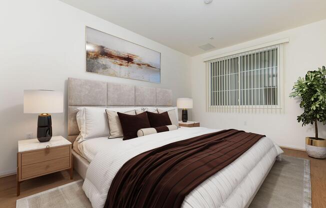 A modern bedroom featuring a large bed with a beige headboard and brown bedding. There are two bedside tables with lamps, and a decorative plant in one corner. A piece of abstract art hangs above the bed, and the room has large windows with vertical blinds. The flooring is light-colored wood, enhancing the bright atmosphere.