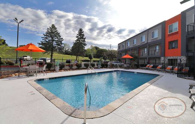 A swimming pool with lounge chairs and tables with orange umbrellas in front of an apartment building.