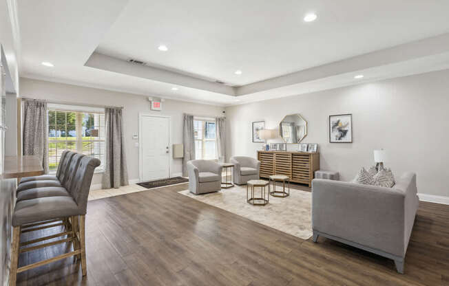 A well-lit, spacious living room with a grey sofa, wooden chairs, and a rug.