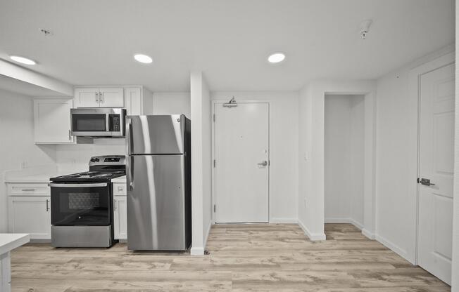 A modern kitchen area featuring a stainless steel refrigerator and microwave, a stove with an oven, and white cabinetry. The space has light-colored hardwood flooring and bright overhead lighting, with a door leading outside visible in the background. The overall design is clean and minimalistic.
