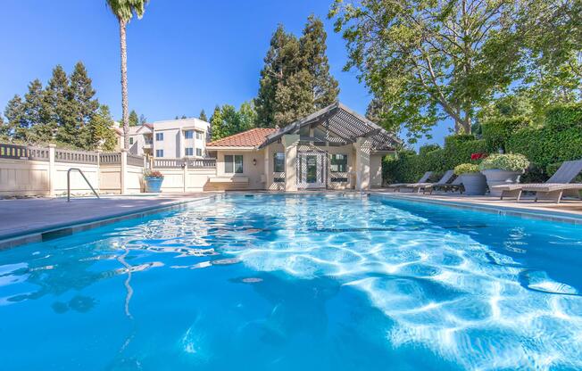 A clear blue swimming pool with a tiled edge, surrounded by lounge chairs and palm trees. In the background, a clubhouse with a tiled roof is visible, along with lush greenery and residential buildings. The sky is bright blue, creating a serene outdoor atmosphere.