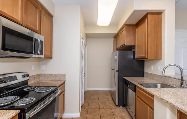A modern, well-lit kitchen featuring wooden cabinets, stainless steel appliances, and granite countertops. The kitchen includes an oven, microwave, refrigerator, and dishwasher, with a tile floor and a spacious layout.