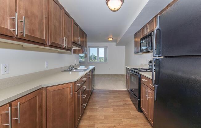A spacious kitchen featuring dark wood cabinets, a black refrigerator, and a black stove. The countertops are light-colored, and there are dual sinks. Natural light comes from a nearby window, providing a view of the outdoors. The flooring is a warm brown, complementing the kitchen's overall design.