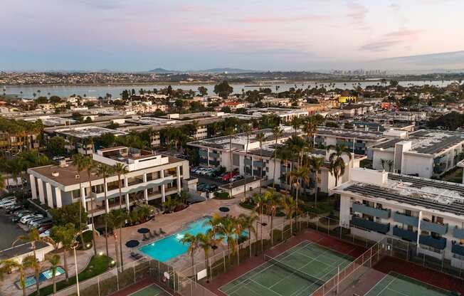 A tennis court is surrounded by a pool and palm trees in a residential area.