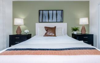 A neatly arranged bedroom featuring a white bed with a textured throw blanket and a large brown pillow. There are two nightstands with lamps, potted plants, and a small flower vase. A piece of wall art with a nature theme hangs above the headboard against a green wall.