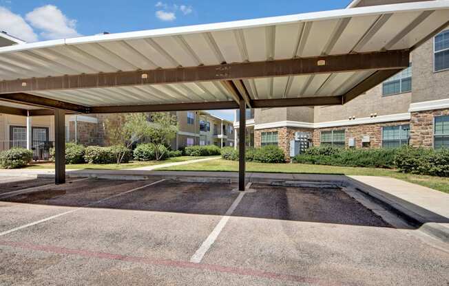 A covered parking area here at Mission Green featuring shaded carport spaces with metal roofing, clearly marked parking stalls, and a paved driveway, with landscaped greenery, walkways, and apartment buildings visible in the background under a bright blue sky.