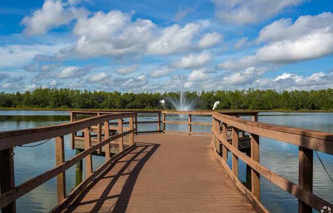 A wooden pier extends into a body of water with a fountain in the distance.