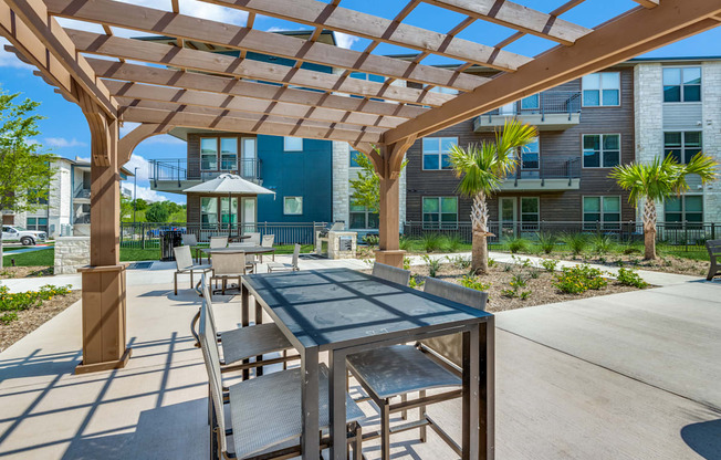 A wooden pergola with a table and chairs is in the foreground of a sunny outdoor area.