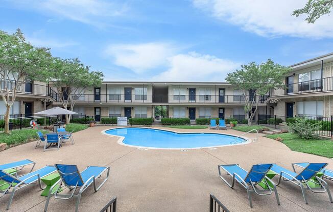 A tranquil courtyard featuring a swimming pool surrounded by lounge chairs. Green shrubs and trees provide shade, while adjacent to the pool, there are umbrellas for relaxation. Two-story building units encircle the pool area, creating a serene atmosphere. Bright, clear sky overhead adds to the inviting scene.