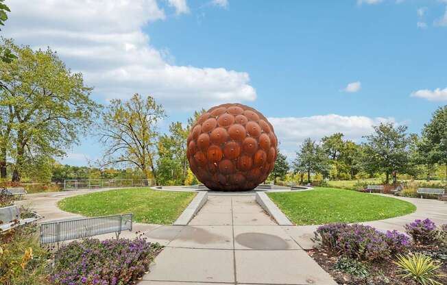 A large orange sphere sculpture is the centerpiece of a park with a walkway leading to it.