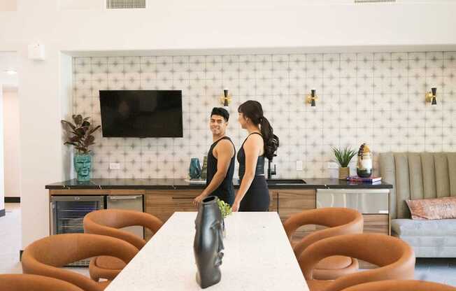 Two people standing in a kitchen with a white table and orange chairs.