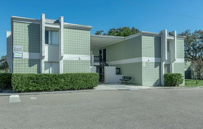A two-story green apartment building with white accents, featuring a covered entrance, stairway, and small bench in front. The surrounding area includes neatly trimmed bushes and a clear blue sky. The building has a modern design and is situated on a quiet street.