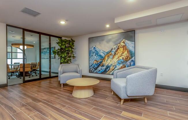 A modern office space featuring two grey armchairs and a round wooden coffee table. A large painting of snow-capped mountains is mounted on the wall, and there is a green indoor plant beside the seating area. Glass walls can be seen in the background, possibly leading to a conference room.