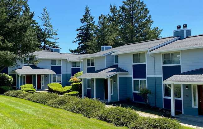 Townhome exterior with blue accents, well-manicured grass, green hedges, patios, and large trees behind. at Hawks Prairie, Olympia, Washington