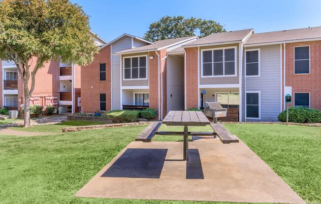 A landscaped courtyard featuring a picnic table on a concrete pad, surrounded by green grass and apartment buildings with screened porches. The buildings exhibit a mix of brick and siding exteriors, with trees providing shade in the area. The scene is bright and inviting under clear blue skies.