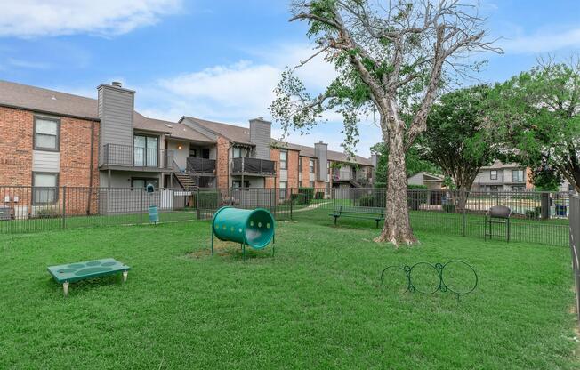 A spacious green pet area featuring grassy lawns, a dog play tunnel, and various benches. In the background, there are two-story apartment buildings with brick exteriors and balconies, surrounded by a fence. Trees provide shade in the area, creating a welcoming environment for pets and residents.