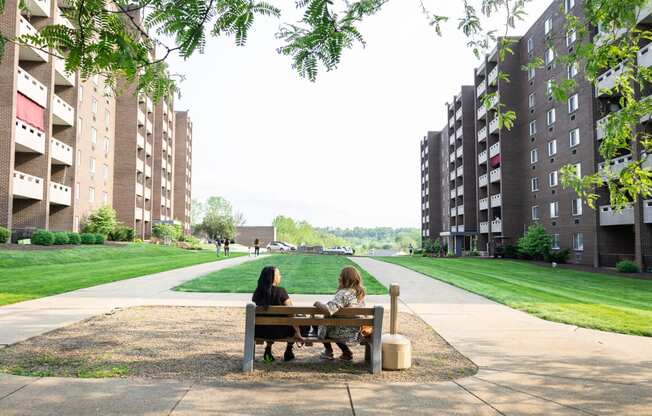 two women sit on a bench in the middle of a grassy area between two apartment buildings