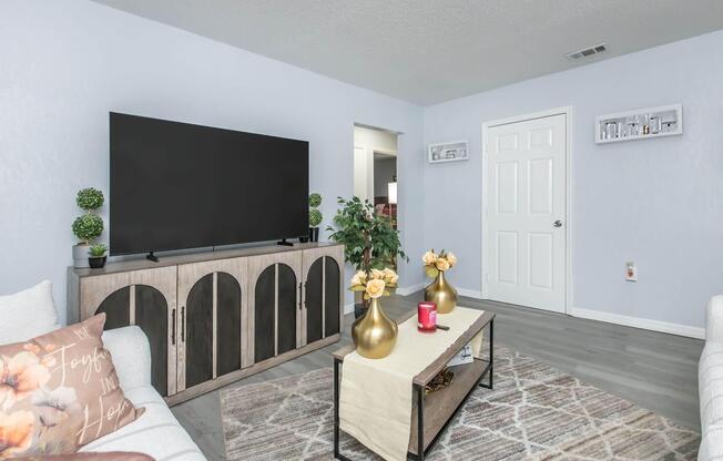 A modern living room featuring a large flat-screen TV mounted on a light blue wall. There is a wooden TV stand with decorative elements, including green plants and gold vases with yellow flowers. A coffee table with a neutral table runner sits in the center, complemented by a red candle. Light-colored sofas complete the inviting space.