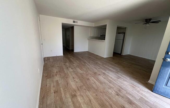 A spacious, empty living area featuring light-colored walls and wood-like flooring. To the right, a doorway leads to a kitchen with white cabinets and appliances. Natural light is coming through a nearby door, which is painted blue. The overall atmosphere is bright and clean.