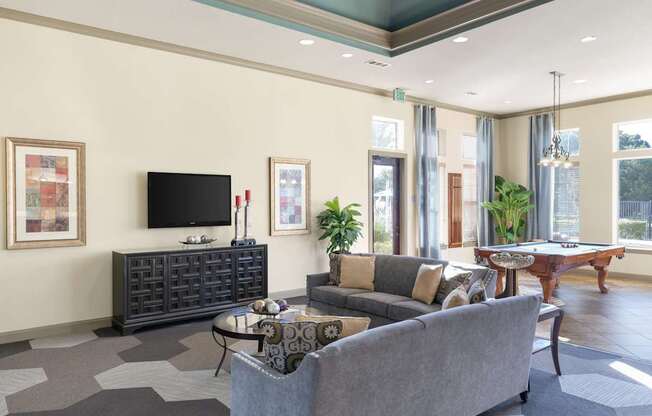 A clubhouse lounge here at Landing at Mansfield with gray sofas and patterned pillows, round glass coffee table, dark media console with wall-mounted TV, framed art, tall windows with drapes, and a pool table near the windowed game area.