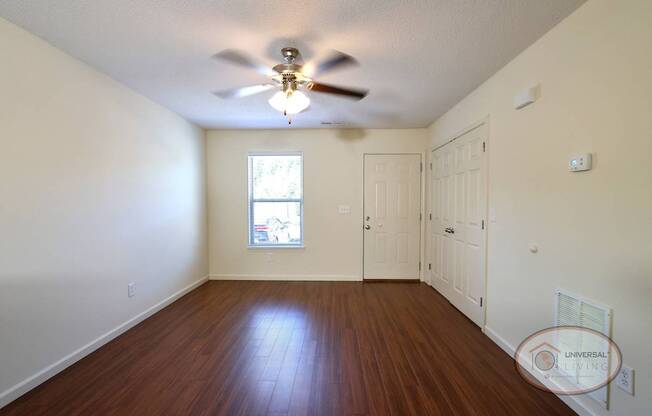 An empty living room with hardwood floors, white walls, a ceiling fan, and a large window.