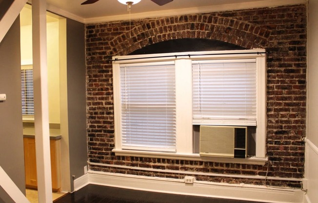 Living room in brookmore apartments in Pasadena featuring an exposed brick wall, large windows with shades, and a window air conditioning unit.