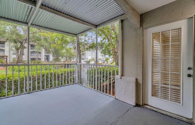 A screened-in porch with a gray floor, featuring a white door with shutters, surrounded by trees and greenery. The space is well-lit with natural light coming through the screens, providing a peaceful outdoor view.