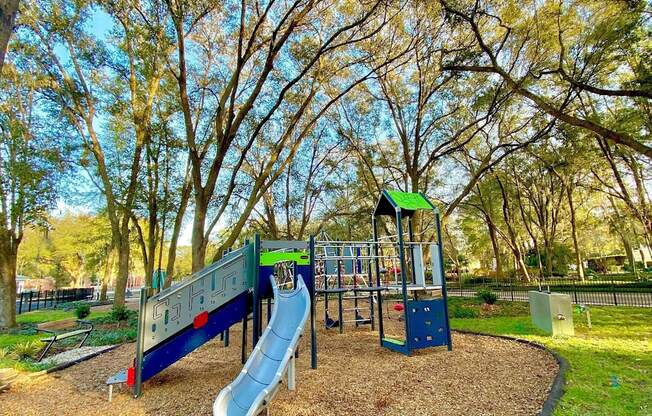 A children's playground on soft wood chips surrounded by grass and large trees near The Flats at Tioga Town Center apartments.