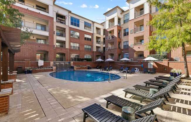 A large outdoor pool area with sun loungers and umbrellas in front of apartment buildings.
