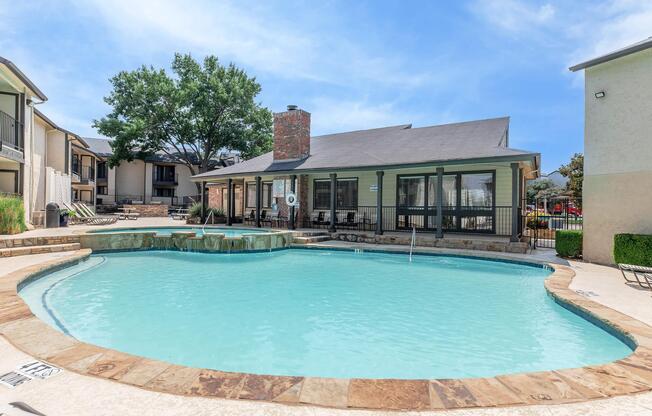 A sparkling outdoor swimming pool with a stone border, surrounded by lounge chairs. In the background, there are two buildings with balconies, and a shaded area with trees. The sky is clear and blue, providing a bright, sunny atmosphere.