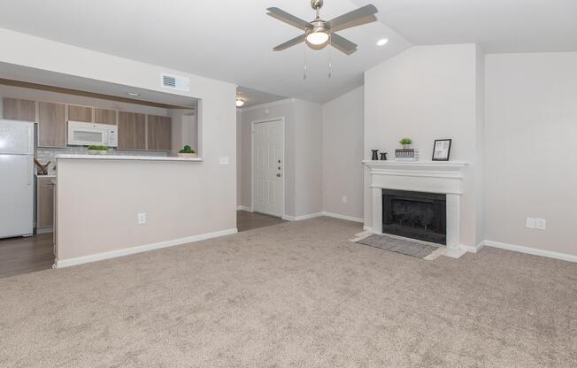 A spacious living area featuring beige walls and carpet. There is a ceiling fan, a white fireplace with a gray mantle, and an open kitchen area with wooden cabinetry. A door leads to the entrance, with a small decorative shelf on the side. Natural light fills the room, creating an inviting atmosphere.