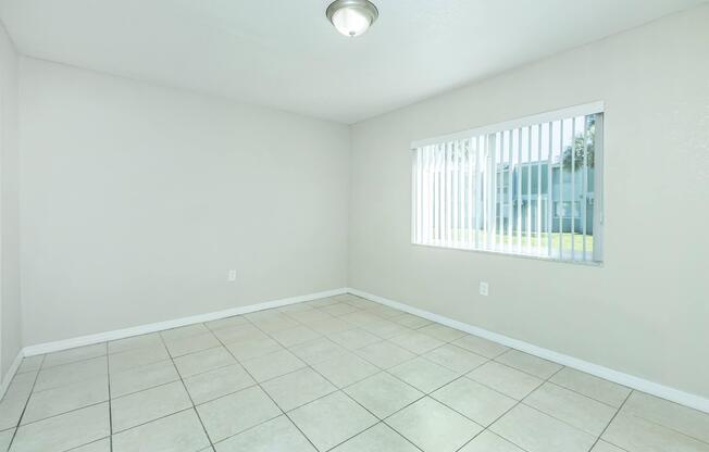 A bright, empty room with light-colored walls and tiled flooring. There is a single light fixture on the ceiling and a window with vertical blinds allowing natural light to enter. The view outside shows greenery, enhancing the room's airy feel.