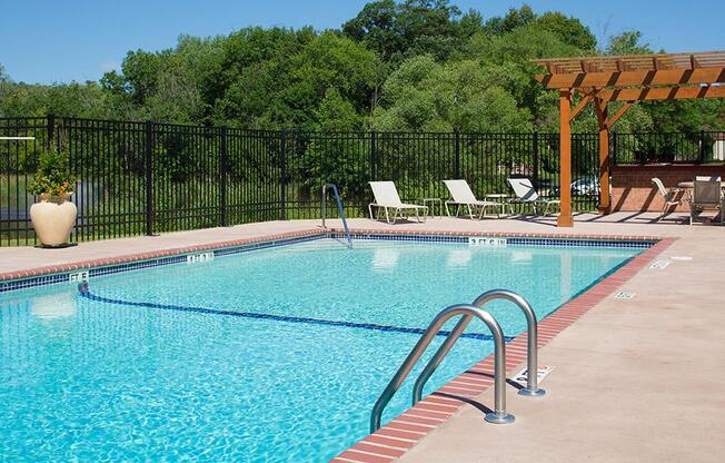 Outdoor pool surrounded by pool chairs and a fence, trees is the background