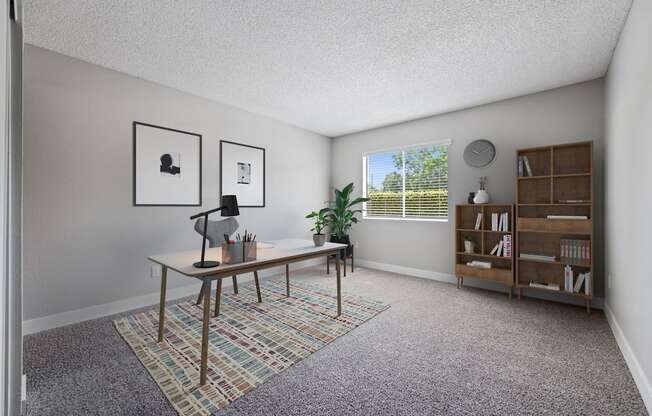 Apartments In Colton, CA - Ardella At Reche Canyon - Home Office With Carpet Flooring, Spacious Desk With Lamp, Chair, Potted Plants, Bookshelves, And Window With Blinds.