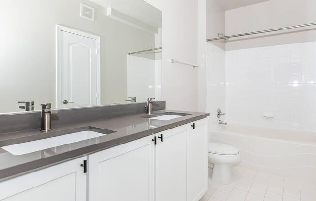 Modern bathroom featuring a double vanity with two sinks and sleek silver faucets, a large mirror above, and a light-colored tiled floor. The adjacent area includes a bathtub and a toilet. The walls are painted in a soft neutral color, creating a clean and spacious aesthetic.
