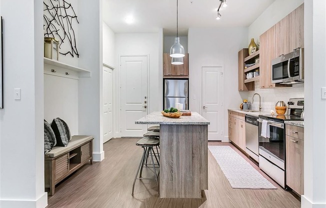 Apartments for Rent in Rowlett, TX - Village of Rowlett - Kitchen with Wood-Style Flooring, Granite Countertops, Wood-Tone Cabinetry, Stainless Steel Appliances, Kitchen Island/Breakfast Bar, Bar-Style Stools, Pendant Lights, Pantry, Storage Shelves, and Bench Seat.