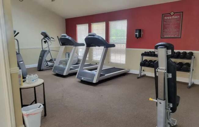 A well-equipped fitness room here at Trinity Place with treadmills, an elliptical machine, and a strength training station. A rack of free weights lines the wall, while large windows with blinds allow natural light into the space. Neutral carpeting, accent walls, and ceiling fans create a comfortable workout environment for residents.