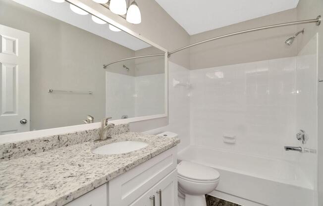 A modern bathroom featuring a white bathtub with a glass shower rod, a beige wall, and a granite countertop with a sink. There are three overhead light fixtures illuminating the space, along with a neatly styled mirror. A closed door is visible to the left. The floor has a wood-like appearance.