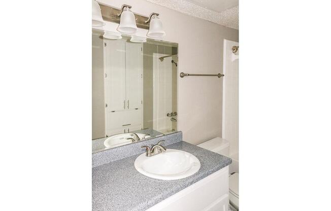 A clean and modern bathroom featuring a light gray countertop with a white sink, a large mirror above, and three light fixtures. The space includes a white toilet and a shower area with a glass door. Storage cabinets are visible in the background, creating a functional layout.