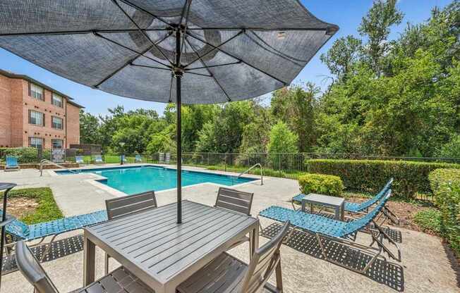 A table with chairs and an umbrella is in the foreground of a pool area.