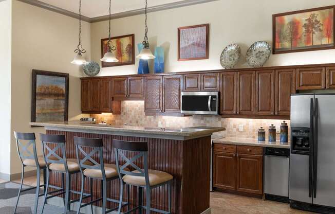 A clubhouse kitchen and bar area here at Landing at Mansfield with dark wood cabinets, long counter with four gray barstools, pendant lights, stainless appliances, tan tile backsplash, and framed colorful wall art above the cabinetry.