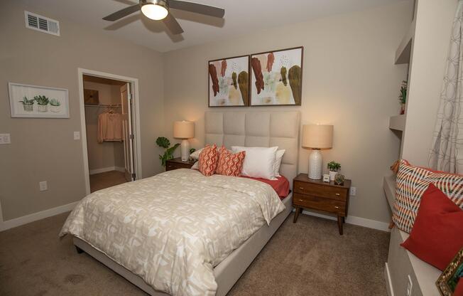 A cozy bedroom featuring a beige bedspread, decorative pillows, and a headboard. There's a nightstand on either side with lamps, and artwork above the bed. A closet is visible in the background, along with a potted plant and some decorative accents on the shelves. Soft lighting enhances the inviting atmosphere.